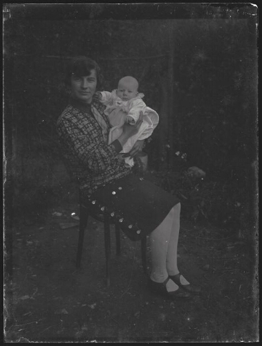 Portrait en pied d'une jeune femme assise sur une chaise tenant un nourrisson dans ses bras vêtu d'une robe de baptême