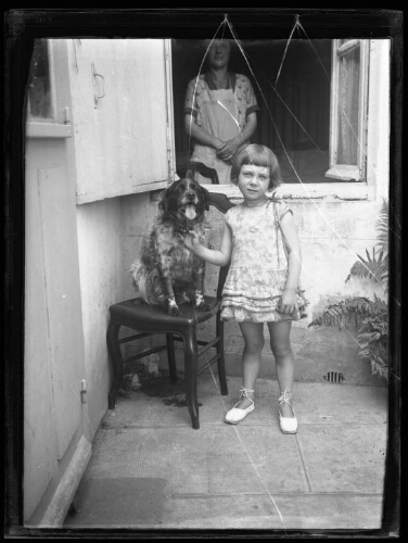 Portrait en pied d'une fillette debout devant une maison tenant par le collier un chien assis sur une chaise près d'elle ; une femme se trouve derrière elle dans l'encadrement d'une fenêtre ouverte