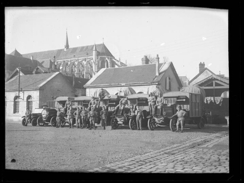 Militaires : des hommes en habit militaire posent devant des camions alignés ; certain d'entre eux sont assis sur les camions ; maisons et église en arrière-plan