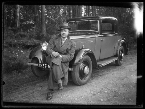 Portrait d'un homme assis sur le par choc avant d'une voiture Peugeot 201 cabriolet, les jambes croisées, vêtu d'un manteau et d'un chapeau panama ; la voiture est immatriculée "1537-MX2"
