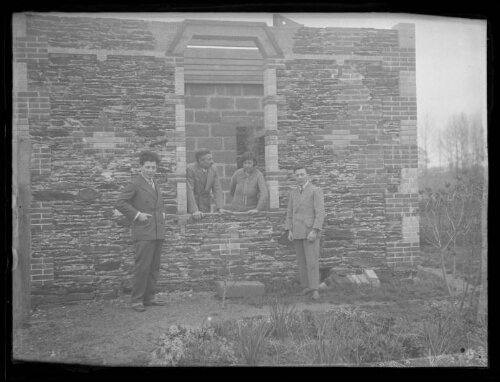 Groupe de personnes : un couple et deux hommes devant une maison en construction ; le couple est à l'intérieur de la maison, en appui sur la fenêtre ; mur en parpaing en arrière-plan dans la maison