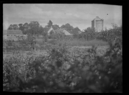 Paysage de campagne : un homme pose dans son jardin une pelle à la main ; maisons, arbres et silo à grain en arrière-plan