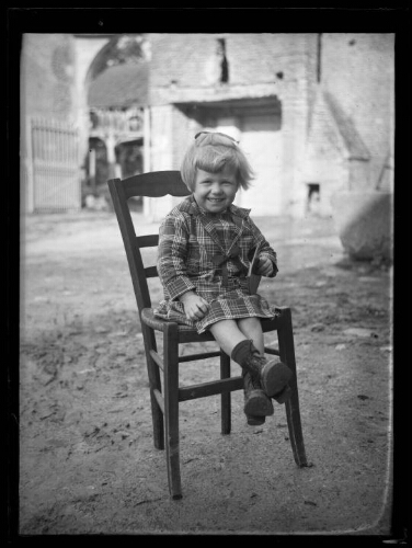 Portrait en pied d'une fillette assise sur une chaise dehors ; elle tient à la main un cahier