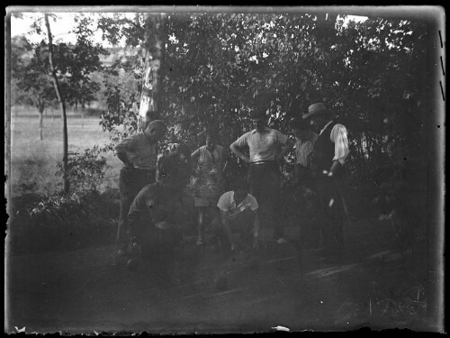 Groupe de personnes : sept hommes et une femme jouent aux boules dehors ; l'un des hommes mesure un point ; arbres en arrière-plan