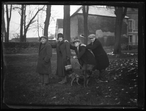 Mise en scène : une femme et trois hommes avec un chien sont debout dehors et se tiennent d'une main par l'épaule ; mur d'enceinte de propriété et maisons en arrière-plan