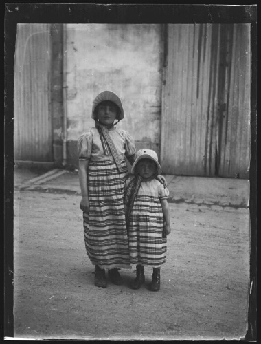 Portrait en pied de deux jeunes filles vêtues à l'identique devant une maison : toutes les deux portent une robe rayée de style "Empire" à manche courte bouffante et un chapeau de style "second Empire" noué autour du cou