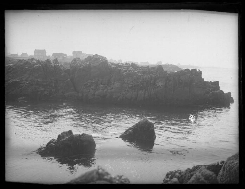 Paysage cotier : deux rochers émergent de l'eau ; des rochers bordent la mer ; quelques maisons en arrière plan
