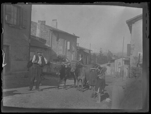 Puy-en-Velay (Haute-Loire), scène rurale : un paysan vêtu d'un chapeau et tenant un baton avec deux enfants se protégeant du soleil avec leur avant-bras encadrent deux vaches tirant une carriole dans une rue de village ; rocher et chapelle Saint-Michel-d'Aiguilhe de Puy-en-Velay en arrière-plan