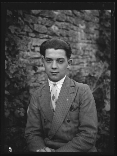 Portrait en buste d'un jeune homme assis dehors devant un mur, vêtu d'une veste à carreaux avec une pochette, d'une chemise et d'une cravate imprimée de losanges