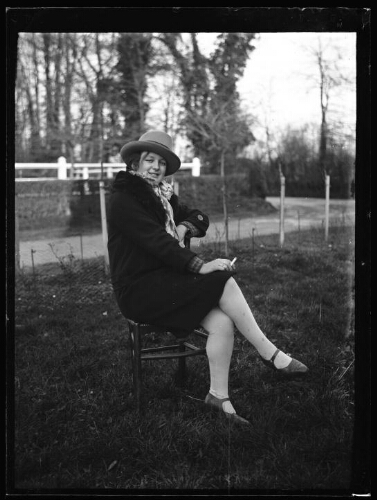 Portrait en pied d'une jeune femme assise dehors sur une chaise dehors ; elle porte un chapeau et tient une cigarette dans sa main