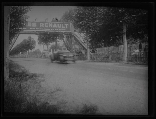 Le Mans (Sarthe) : circuit des 24 heures : voiture sur le circuit au niveau d'une passerelle où un panneau publicitaire "Huiles Renault... meilleures" est accroché ; spectateurs derrière une palissade en bois en arrière-plan