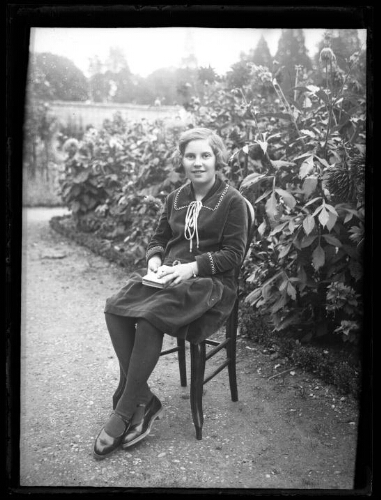 Portrait en pied d'une femme assise dehors sur une chaise dans une allée de jardin, avec un livre fermé sur ses genoux