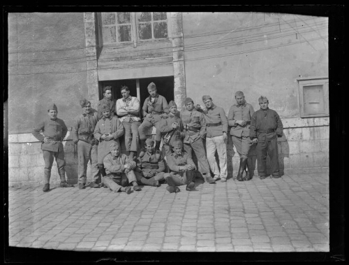 Militaires : quatorze hommes en habit militaire posent alignés devant un bâtiment ; les trois hommes devant sont assis par terre, les trois hommes derrière sont assis sur un muret ; l'un des militaires debout tient une trompette