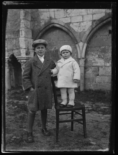 Portrait en pied de deux enfants debout devant un monument : le plus jeune est debout sur une chaise