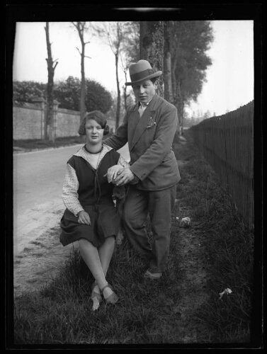 Portrait en pied d'un couple au bord d'une route ; la femme est assise, l'homme est debout près d'elle