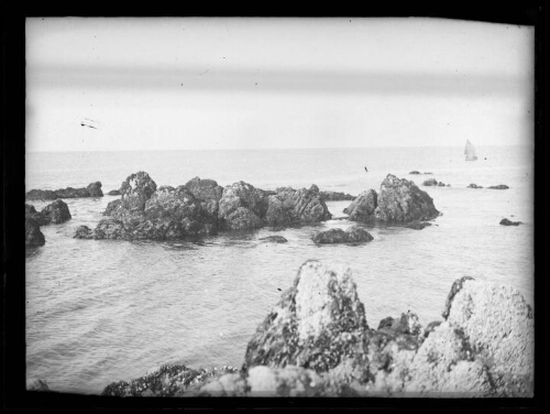 Paysage côtier : rochers émergeant de la mer ; voilier en arrière-plan