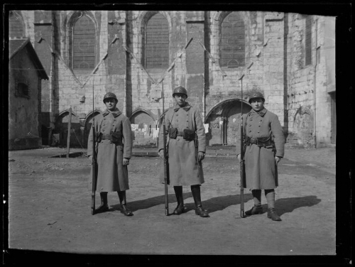 Militaires : trois hommes en habit militaire posent debout devant une église ; ils sont vêtus de manteaux, portent un casque et tiennent chacun une baïonnette