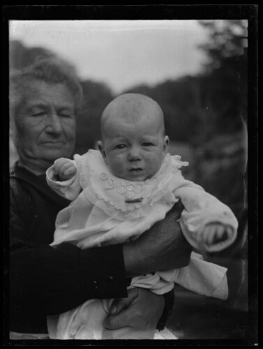 Portrait d'un nourrisson dans les bras d'une femme âgée ; il est vêtu d'une robe de baptême avec un col en dentelle brodée