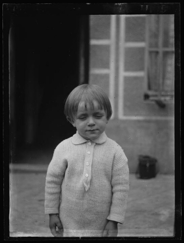 Portrait, en buste, d'un jeune garçon devant une maison, vêtu d'un pull boutonné à manche longue ; ses yeux sont fermés