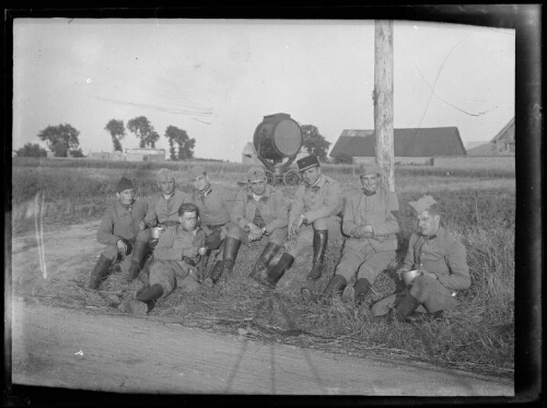 Militaires : huit hommes en habit militaire sont assis sur le bord d'une route devant un projecteur pour l'atterrissage des avions sur des aérodromes ; l'un d'eux tient une tasse et deux autres leur gamelle ; l'un d'eux porte le képi du 401e régiment d'artillerie anti-aériens ; hangar et maisons en arrière-plan