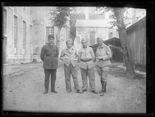 Militaires : quatre hommes en habit militaire posent debout dehors dans la cour d'une bâtisse ; l'un des trois hommes porte un képi du 401e régiment d'artillerie anti-aériens