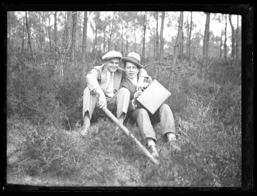 Deux hommes assis dans un bois ; l'un tient une petite valise, l'autre un bâton ; arbustes en arrière-plan