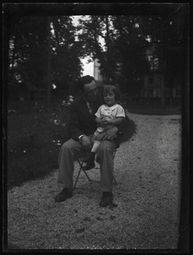 Portrait en pied d'un homme assis dehors sur une chaise de jardin avec une fillette sur ses genoux ; l'homme regarde l'enfant