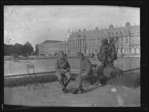 Versailles (Yvelines) : deux hommes en habit militaire assis sur des chaises de jardin près d'une statue d'angelots du parc du château de Versailles ; château de Versailles en arrière-plan