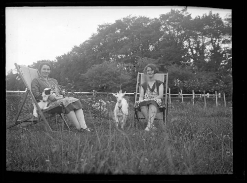 Portrait de deux femmes assises dans des transats dans un pré ; l'une d'elle tient un chien sur ses genoux, l'autre tient une chèvre avec une chaine
