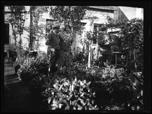 Groupe de personnes : un couple et un homme avec un un chat dans ses bras posent devant une maison au jardin fleuri