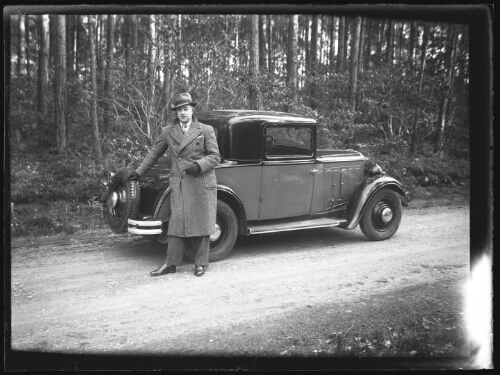 Portrait d'un homme debout près d'une voiture Peugeot 201 cabriolet, vêtu d'un manteau, d'un chapeau panama et de gants ; il tient une cigarette dans une main, son autre main est posée sur la roue de secours de la voiture