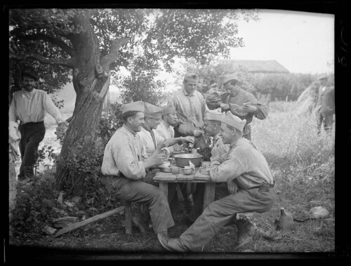 Militaires : cinq hommes en habit militaire assis autour d'une table en bois installée dans un champ se restaurent ; un récipient et des tasses sont posés sur la table