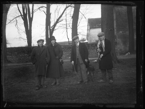 Groupe de personnes : une femme et trois hommes avec un chien posent debout dehors ; mur d'enceinte de propriété et maisons en arrière-plan