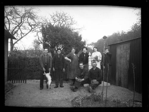 Portrait de groupe : Marc Bry avec des amis, deux femmes et cinq hommes dans une cour près d'un cabanon ; deux des hommes sont assis sur le socle d'une pompe à puit