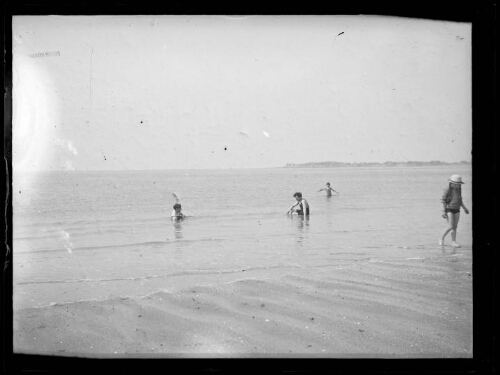 La Baule (Loire-Atlantique) ? : deux hommes sont assis dans la mer face-à-face, un enfant se baigne derrière eux et une fillette avec des tresses et un chapeau se dirige vers la plage