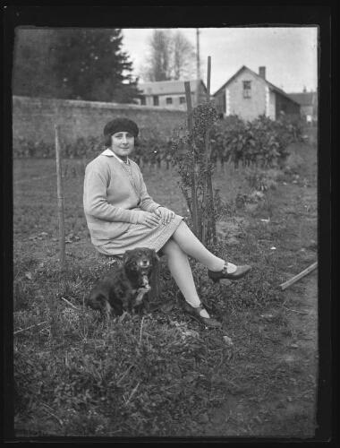 Portrait en pied d'une jeune femme assise de trois quart dehors sur une chaise dans un jardin, avec un chien assis près d'elle