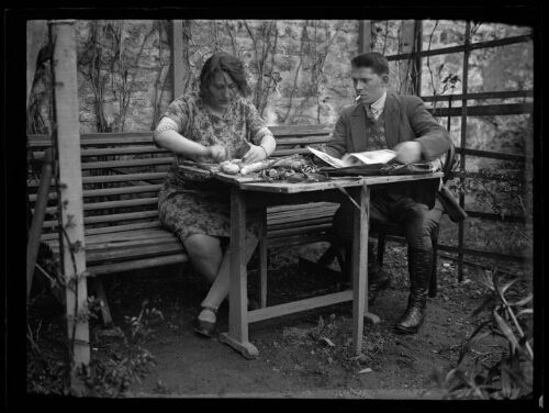 Scène de la vie quotidienne : un couple est assis à une table sous une pergola ; la femme épluche des pommes de terre et l'homme feuillette un magazine, une cigarette à la bouche