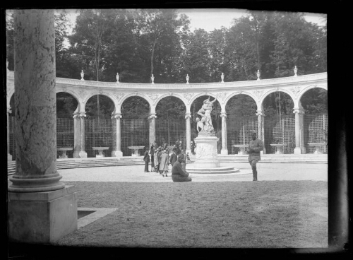 Versailles (Yvelines) : bosquet de la Colonnade, jardins du château de Versailles ; deux hommes en habit militaire posent devant la statue, l'un debout, l'autre assis, et un groupe de personnes, derrière eux, observent la statue