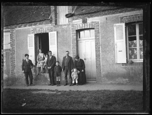 Groupe de personnes : deux femmes, trois hommes et trois enfants devant une maison ; l'une des femmes est assise sur le rebord de la fenêtre ouverte avec un enfant
