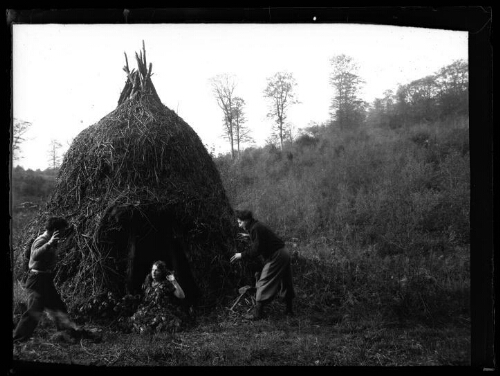 Marc Bry avec un couple d'amis près d'une meule de foin transformée en cabane : les deux hommes brandissent les bras dans l'intention d'effrayer ou d'attraper la femme recouverte de feuilles, le 4 mai 1936