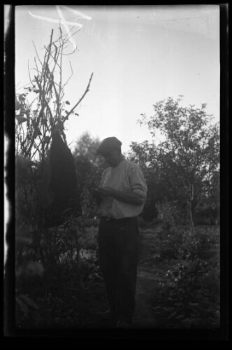 Portrait en pied d'un homme dans son jardin ; sa veste est accroché dans un arbuste près de lui