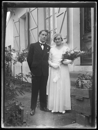 Mariage : un couple de mariés pose debout devant les marches d'entrée d'une maison ; la mariée porte un voile et tient un bouquet de fleurs dans ses mains