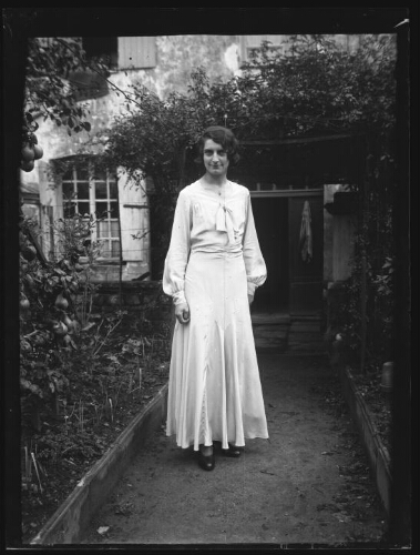 Portrait en pied d'une femme debout dans l'allée menant à l'entrée d'une maison ; elle est vêtue d'une robe longue fermée sur la poitrine par un ruban noué et d'un collier (robe de mariée ou personne invitée à un mariage ?)
