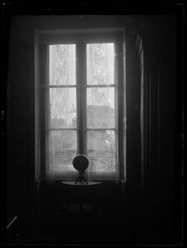 Alençon (Orne) : fenêtre fermée aux rideaux brodés de fleurs et aux volets ouverts ; un globe terrestre est posé sur un guéridon devant la fenêtre ; maison d'Alençon visible à travers la fenêtre