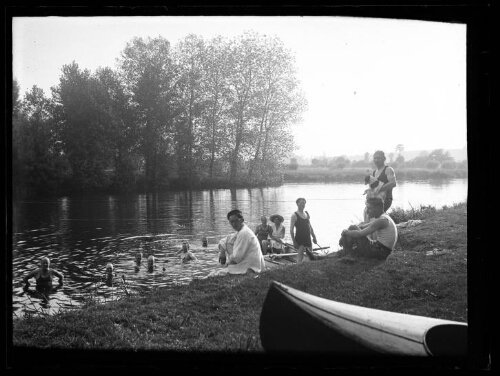 Scène de baignade : six personnes se baignent dans une rivière, trois autres sont dans une barque près d'eux, deux sont assis sur la berge et un homme est debout avec un chien dans les bras ; extrémité d'un kayak au premier plan ; arbres en arrière-plan