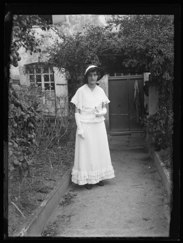 Portrait en pied d'une femme debout dans l'allée menant à l'entrée d'une maison ; elle est vêtue d'une robe longue avec des volants, d'un châle, de gants et d'un chapeau (robe de mariée ou personne invitée à un mariage ?)