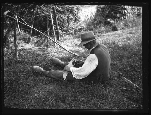 Gustave Chevalier, le grand-père de Marc Bry, assis dans l'herbe, bat sa faux à herbe avec un marteau au lieu-dit « Le Gué » à Essay (Orne) le 6 juillet 1930