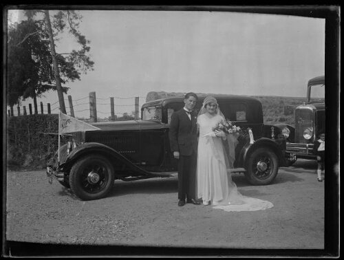 Mariage : un couple de mariés pose debout devant une voiture Renault ; la mariée porte un voile et tient un bouquet de fleurs dans sa main