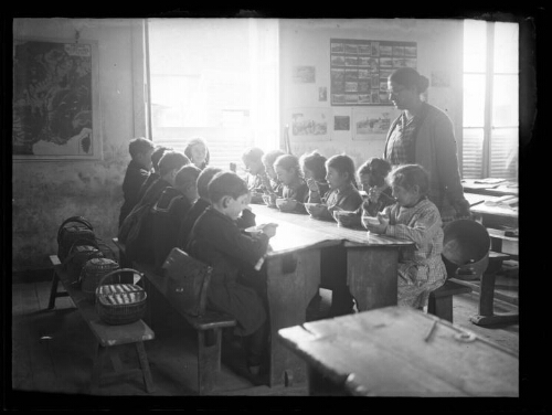 Scène d'écoliers : les élèves, assis dans une salle de classe, ont chacun un bol et mangent ; l'institutrice est debout près d'eux avec un faitout à la main
