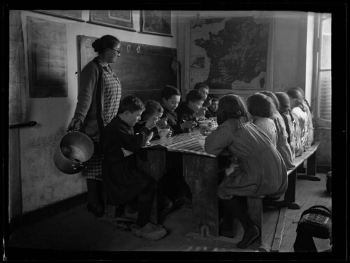 Scène d'écoliers : les élèves, assis dans la salle de classe, ont chacun un bol et mangent ; l'institutrice est debout près d'eux avec un faitout à la main ; les garçons sont assis d'un côté de la table, les filles de l'autre